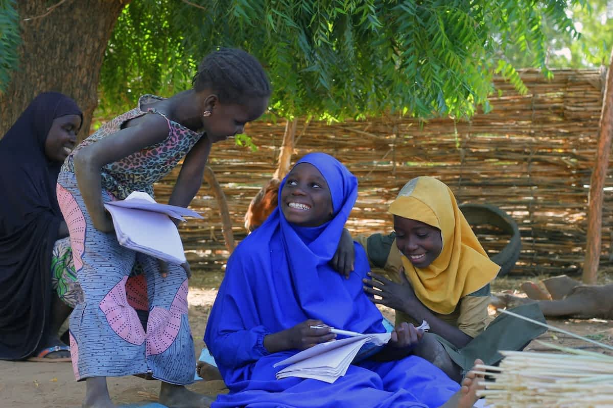 Tre piger laver lektier i skyggen af et træ. De smiler til hinanden. Niger