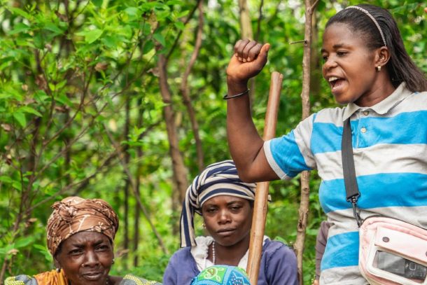 tre kvinder i Afrika. To sidder ned og den tredje står på og har den ene hånd i vejret